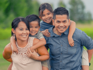 Happy family of four smiling outdoors, with children on parents' backs.