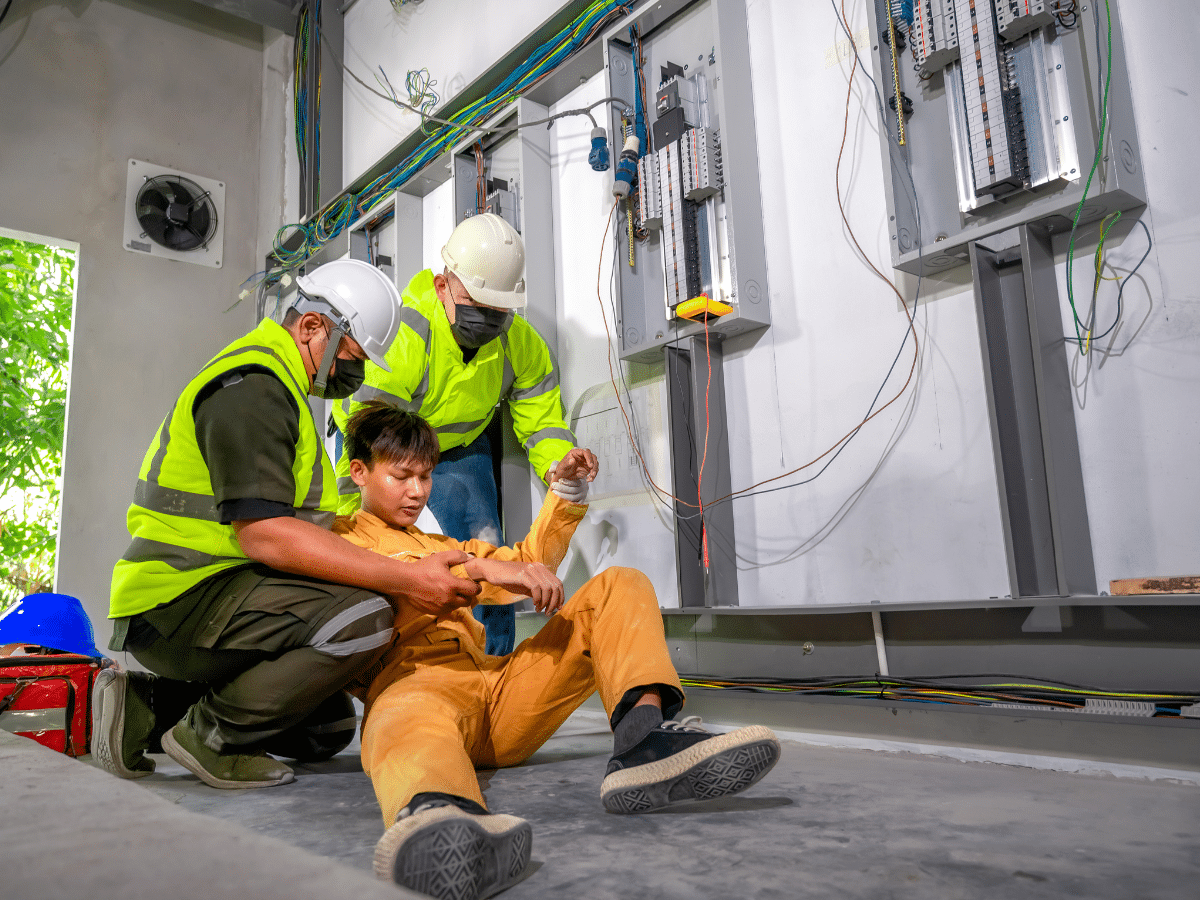 Two electricians in safety gear assist an injured coworker on the floor near electrical panels. 