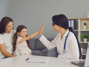  smiling doctor high-fiving a young girl sitting with her mother during a consultation.