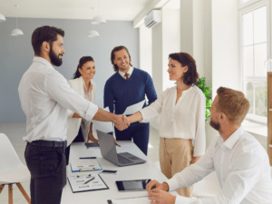  A group of business professionals in an office setting smiling as two individuals shake hands across a conference table.