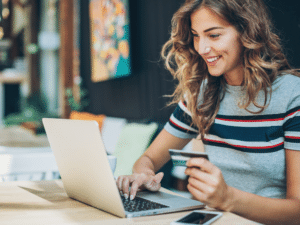 Young woman sitting at a table, smiling while making an online purchase on her laptop with a credit card in hand.